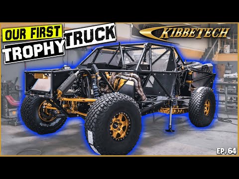 A partially built off-road trophy truck in a workshop with visible roll cage, tires, and suspension components, large text says our first trophy truck KibbeTech in the background, episode 64 displayed at the bottom corner.