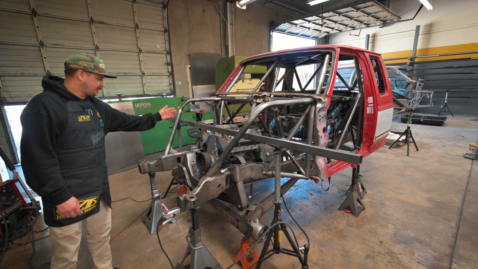 A person in a workshop is working on the chassis of a prerunner truck with an exposed metal framework. The red cab of the truck is attached to the frame, and various tools and equipment are visible in the surroundings.