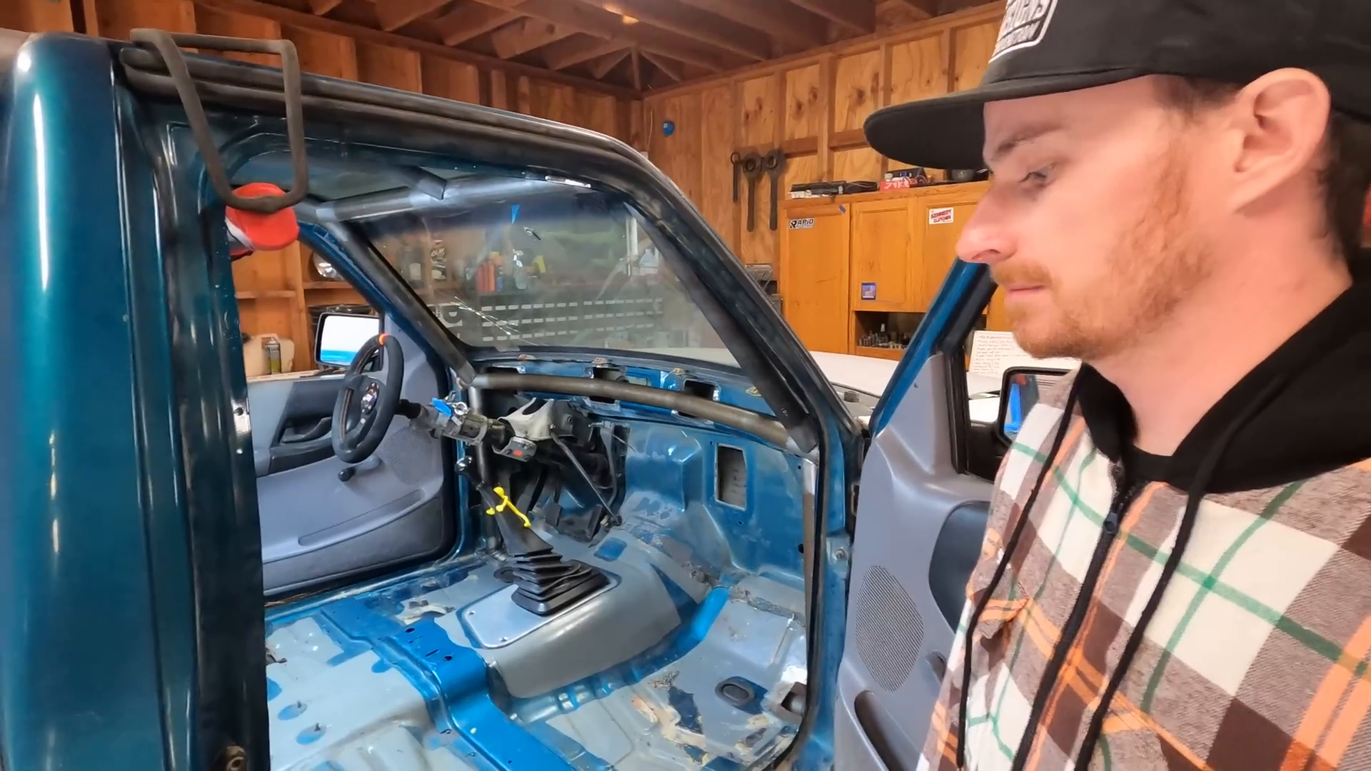 A person stands next to a blue vehicle in a garage with the driver's door open. The vehicle's interior is stripped, showing the bare metal floor and a partially disassembled dashboard. Tools and equipment are visible in the background.